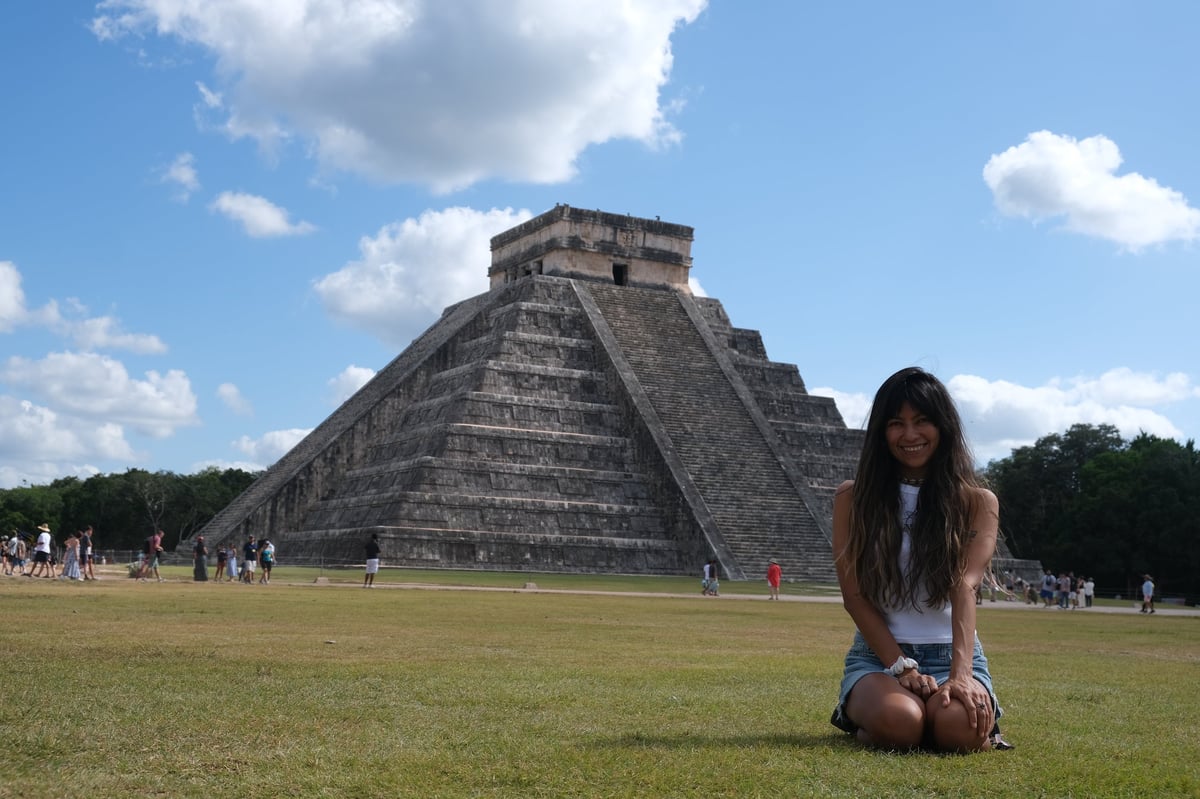 Woman kneeling on grass in front of the Pyramid of Kukulkan at Chichen Itza archaeological site with blue sky and clouds behind