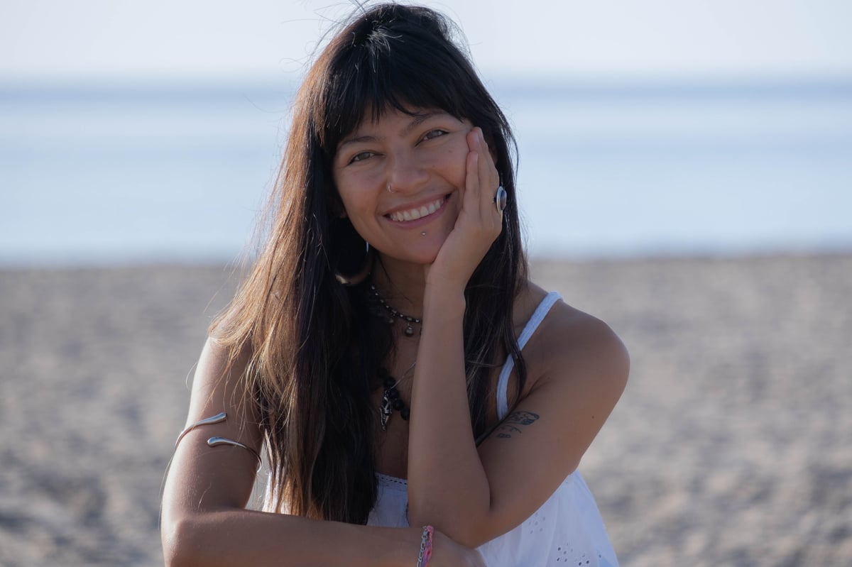 Woman with long dark hair smiling at the beach during sunset, wearing a white tank top and necklace